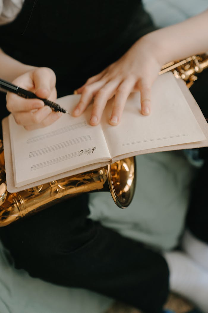 Person writing music in a notebook while holding a saxophone, capturing creativity.